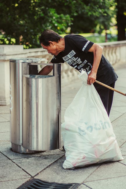 A man with short dark hair, wearing a black T-shirt with white text and dark trousers, is seen using a long-handled tool to place a large white rubbish sack into a stainless steel litter bin with dual compartments, situated on a paved walkway. The bin is positioned next to a stone balustrade, with lush green trees and some natural daylight visible in the background. The man appears focused on discarding waste as part of a street cleaning or waste collection activity. The scene reflects an outdoor environment associated with waste management, showcasing the process of rubbish disposal close to a public park or urban space, possibly involving private or independent waste handling services such as those provided by House Clearance Fulham, aligned with alternative rubbish collection methods often used in residential or commercial clearance tasks.