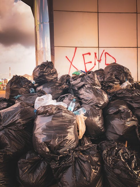 A large pile of black garbage bags, some with visible plastic bottles and packaging, stacked outdoors against a tiled wall with graffiti that reads 'XEND' in red spray paint. The bags are irregularly arranged, with some leaning against the wall and others piled atop each other, indicating an on-site waste clearance or private rubbish collection. The background shows an overcast sky with grey clouds, and the scene suggests a typical rubbish removal situation where waste has been gathered for transport or disposal, a common service provided by companies like House Clearance Fulham. The environment appears urban, with the waste situated on a paved surface adjacent to a modern building, reflecting an area undergoing waste clearing or rubbish removal activities in Fulham, compatible with independent collection or alternative waste handling services.