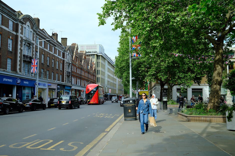 A busy street scene featuring a row of historic brick buildings with retail shops on the ground floor, including a pharmacy, a beauty store, and an office supply shop, all displaying Union Jack flags hanging outside. Several black cars are parked along the curb, with an iconic red double-decker bus traveling down the street. To the right, there is a sidewalk shaded by lush green trees, one of which is prominent in the foreground, with a park planter at its base filled with flowering plants. Two women walking along the pavement are dressed in casual clothing, with one wearing sunglasses and a blue jacket, and the other in a white headscarf and light attire, carrying shopping bags. A black litter bin is positioned near the sidewalk, and a few pedestrians are visible further down the street, some near a small group gathered near the park area. The scene is illuminated by natural daylight, creating a vibrant urban atmosphere typical of central Fulham or similar residential and commercial districts, with a subtle suggestion of alternative waste handling through the well-maintained condition of public spaces, aligning with private rubbish removal services like those offered by House Clearance Fulham.