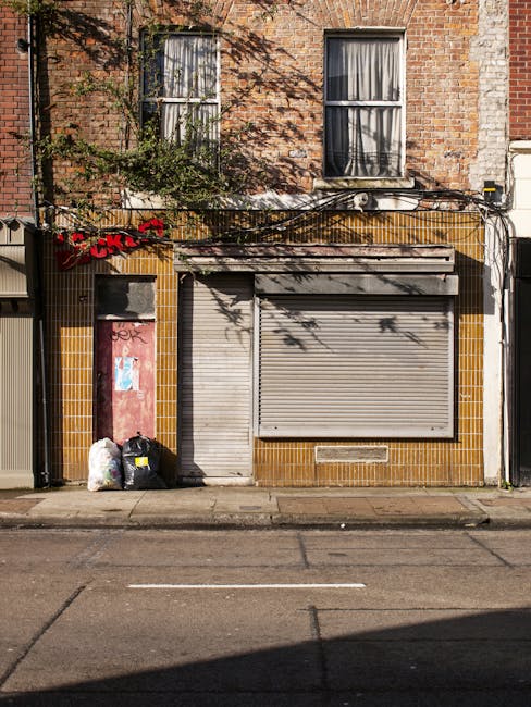 A busy street scene featuring a row of historic brick buildings with retail shops on the ground floor, including a pharmacy, a beauty store, and an office supply shop, all displaying Union Jack flags hanging outside. Several black cars are parked along the curb, with an iconic red double-decker bus traveling down the street. To the right, there is a sidewalk shaded by lush green trees, one of which is prominent in the foreground, with a park planter at its base filled with flowering plants. Two women walking along the pavement are dressed in casual clothing, with one wearing sunglasses and a blue jacket, and the other in a white headscarf and light attire, carrying shopping bags. A black litter bin is positioned near the sidewalk, and a few pedestrians are visible further down the street, some near a small group gathered near the park area. The scene is illuminated by natural daylight, creating a vibrant urban atmosphere typical of central Fulham or similar residential and commercial districts, with a subtle suggestion of alternative waste handling through the well-maintained condition of public spaces, aligning with private rubbish removal services like those offered by House Clearance Fulham.