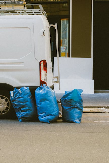The image depicts the rear side of a white commercial van parked on a paved street, adjacent to a building with a glass door and window. Four large blue rubbish bags, made of shiny plastic material, are positioned on the ground near the back of the van. The bags appear to be full, with some showing slight bulges and creases. The van’s rear lights and part of its rear wheel are visible, and the environment suggests an urban setting. Behind the bags, there is a low concrete curb and a narrow strip of sidewalk, with the building's exterior featuring a dark-colored wall and a glass frontage. The scene reflects a typical private rubbish disposal or on-site clearance situation, suited for independent collection services such as those provided by House Clearance Fulham. The overall lighting is natural, with daylight evenly illuminating the scene, emphasizing the practical aspect of rubbish removal and waste management.