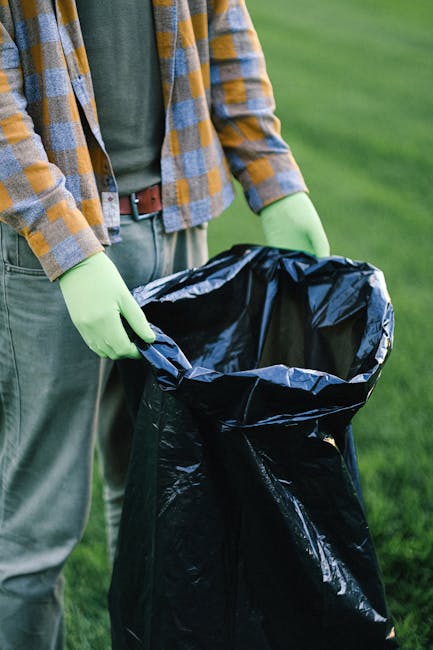 A person standing outdoors on a grassy area is wearing a plaid shirt with shades of yellow, orange, and gray, over a green T-shirt. They are also wearing light gray trousers secured with a brown belt and are equipped with light green disposable gloves. The individual is holding open a large black plastic rubbish bag with both hands, preparing to collect waste. The background features a neatly maintained lawn with vibrant green grass, and the scene is well-lit, suggesting daylight. The image depicts an act of rubbish collection, aligning with services offered by companies like House Clearance Fulham, which handle private waste disposal and on-site clearance operations. The focus is on the manual process of rubbish handling, emphasizing the importance of careful waste collection in a garden or outdoor environment, with the black bag ready for detritus removal or disposal through alternative waste handling methods.