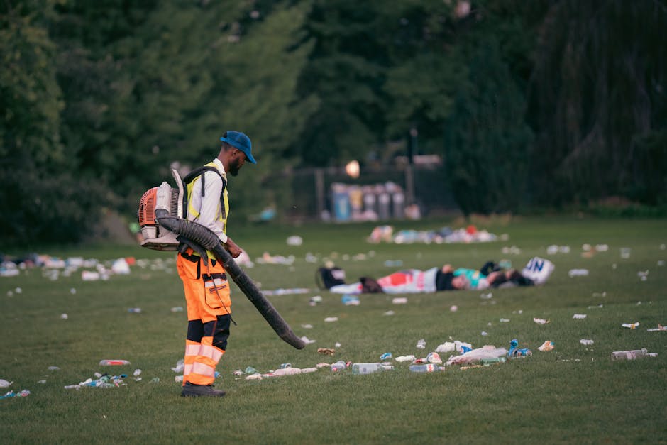 A male worker wearing high-visibility orange trousers with reflective stripes, a white jacket with black accents, and a blue cap is using a leaf blower with an orange and black backpack attachment to clear litter from a grassy park area. The worker is standing on a well-maintained lawn, with a focused posture, holding the blower's nozzle towards the ground. In the background, several individuals are lying on the grass, with litter scattered across the area, including plastic bottles, cans, and paper debris. Behind them, there are tall trees providing shade, and a row of small utility units or storage containers is visible in the distance. The scene is captured in natural daylight, highlighting the contrast between the clean, maintained grass and the scattered rubbish, implying an effort towards environmental cleanliness and waste management services, such as those provided by companies like House Clearance Fulham for private waste disposal and rubbish removal.