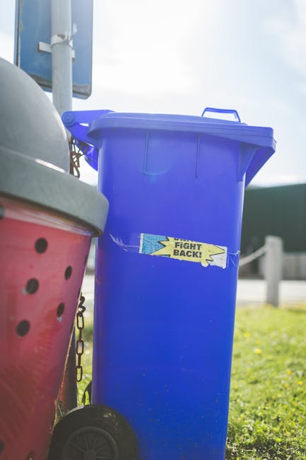 A large blue plastic wheeled rubbish bin with a closed lid, positioned outdoors on a grassy area, with a small rectangular sticker on the front that reads 'Fight Back!' in yellow and black text. To the left of the blue bin, part of a red and blackish-green waste container with ventilation holes and a metallic lid is visible, mounted on a pole with a chain attachment. In the background, there are metal posts, a concrete pavement, and a blurred green structure, suggesting an urban or suburban environment near a roadway or public space. The scene is illuminated by natural daylight, highlighting the textures of the plastic and the grassy surface underneath, which indicates an outdoor waste collection area that could relate to private rubbish disposal or alternative waste handling services, such as those offered by House Clearance Fulham.