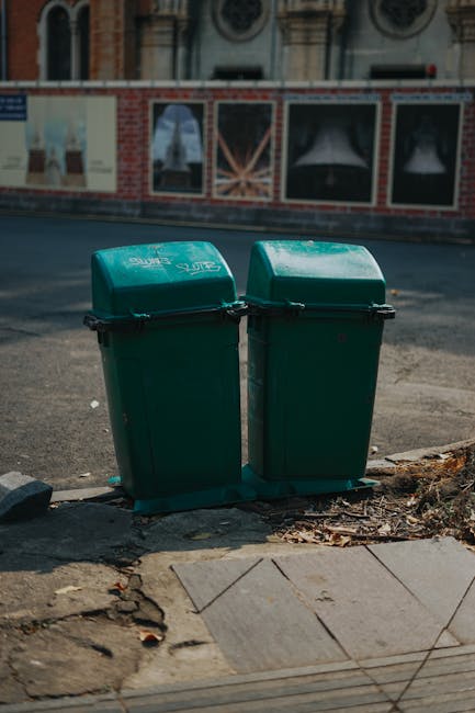 Two green wheeled rubbish bins with closed lids are positioned side by side on a concrete pavement at the edge of a street, in front of a low curb and adjacent to a cracked section of pavement with scattered dry leaves. The bins have a matte finish and appear to be made of durable plastic, with black handles and wheel axles. Behind them, the roadway is visible with a dark, textured asphalt surface. In the background, there is a brick wall with large display windows, featuring images of curtains, and a residential building with ornate architectural details above the wall. The scene is illuminated by natural daylight, casting subtle shadows, and the environment suggests an urban setting suitable for private waste collection or rubbish removal services, such as those offered by House Clearance Fulham. The image subtly evokes the process of independent waste disposal outside a property, highlighting the role of local rubbish collection solutions within Fulham and nearby areas.
