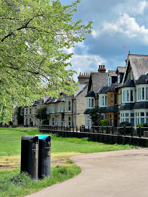 Photograph of a residential street scene featuring a line of traditional Victorian-style terraced houses with bay windows and slate roofs, situated behind a well-maintained grassy verge enclosed by a black metal fence. In the foreground, two black outdoor rubbish bins with blue and black lids are positioned at the edge of a paved pathway. A large leafy tree with fresh green foliage extends over the scene from the left side, providing partial shade and contrast against the partly cloudy sky with patches of blue. The environment appears clean and orderly, indicative of private property maintenance or a managed communal space, complementing the context of rubbish collection or waste management services often associated with residential clearance activities. The natural lighting highlights the textures of the houses' stone facades and the vibrant green of the grass and tree leaves, contributing to the overall tidy and well-kept appearance typical of an area served by independent waste disposal or rubbish removal providers such as House Clearance Fulham.